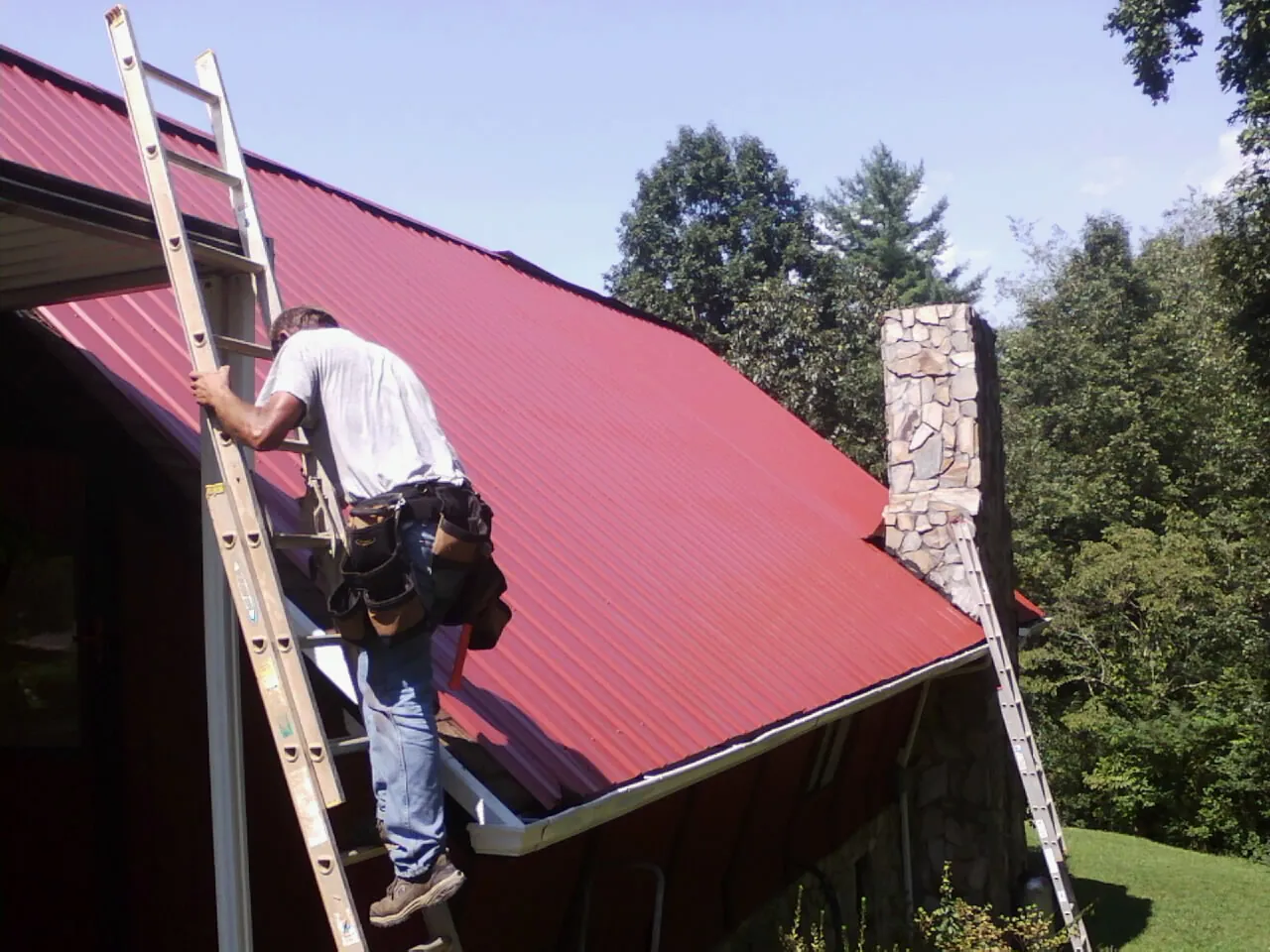 Roofer on a ladder beside a red metal roof and stone chimney — Soffit & Fascia Repair by Roofing Brothers in Pocono Pines
